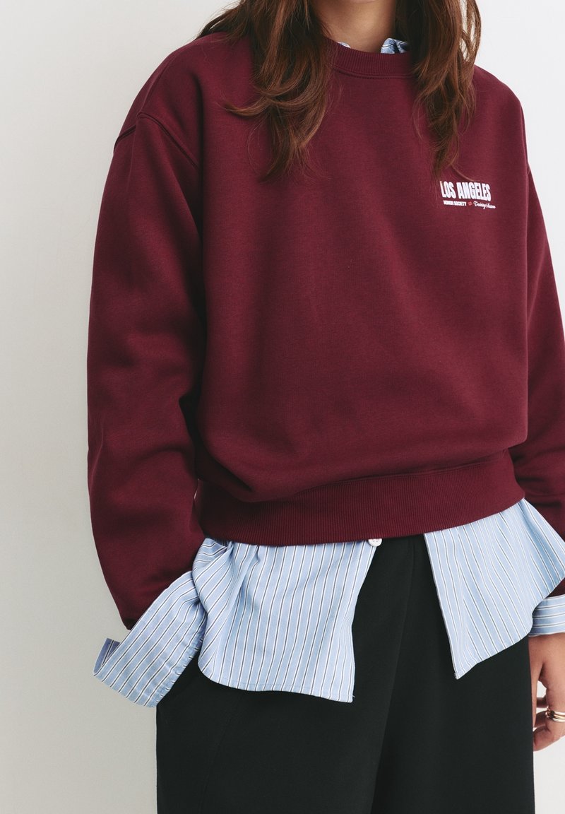 Personne portant un sweat-shirt bordeaux avec le texte "Los Angeles" sur une chemise aux rayures bleues et blanches et un pantalon noir, visible du cou jusqu’aux hanches.