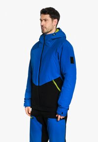 Blaue und schwarze isolierte Jacke mit Kapuze, die grüne Akzente, Reißverschlusstaschen und ein schlankes Design aufweist. Das Material wirkt langlebig und glatt.