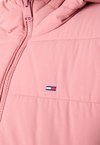 Veste matelassée rose avec fermeture éclair à l'avant et petit logo rectangulaire brodé sur la poitrine en bleu, blanc et rouge.