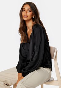 Blusa nera in satin con scollatura a V, spalle plissettate e polsini elasticizzati. Abbinata a pantaloni chiari, situata in un contesto neutro.