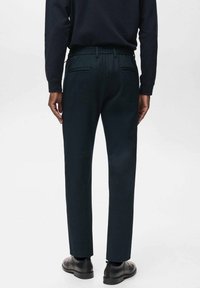 Pantalons vert foncé avec une taille élastique, deux poches arrière, coupe droite et texture lisse. Portés avec une chemise noire à manches longues.