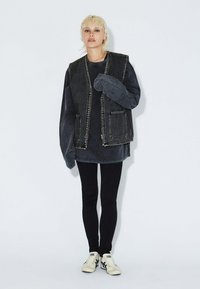 Černá oversized džínová vesta s velkými kapsami, nošená přes tmavý svetr a spárovaná s černými legínami a světlými teniskami.