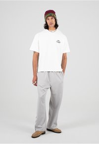 Hvit kortermet skjorte med grafikk, lysegrå vid bukse, beige slip-on sko, og en maroon lue med grønne detaljer.