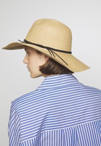 Hut aus Stroh mit breiter Krempe in Beige, mit einem schwarzen geflochtenen Band. Das Modell trägt ein blau-weiß gestreiftes Hemd.