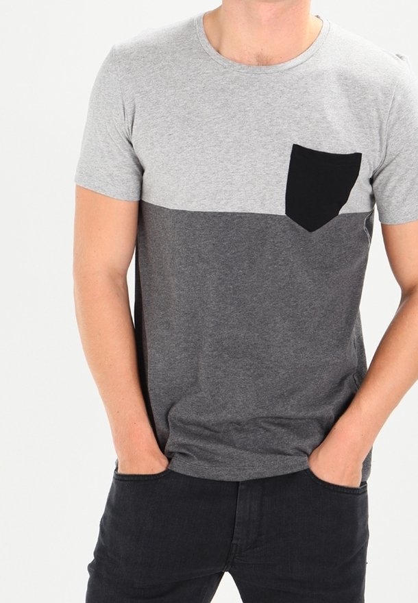 Homme portant un t-shirt gris bicolore à manches courtes avec une poche poitrine noire, les mains dans les poches d'un pantalon noir.