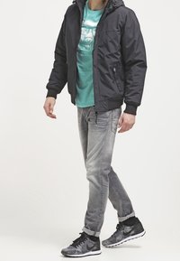 Giacca nera con zip e cappuccio, t-shirt grafica teal, jeans grigi chiari e sneakers nere a fantasia. Vestibilità casual, tessuto morbido e design minimalista.