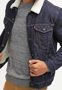 Jeansowa kurtka z kołnierzem z owczej wełny, w ciemnoniebieskim kolorze, z przednimi kieszeniami i jasnymi detalami przeszycia, noszona na szarym swetrze.