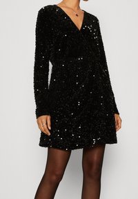 Svart paljettminiklänning med långa ärmar och V-ringning. Tyget är texturerat och glittrande, vilket tillför visuell intresse till designen.