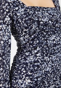 Mörkblå blommig klänning med långa ärmar, prydd med mönster av vita och ljusblå blommor. Tyget är rynkat vilket skapar en texturerad detalj.