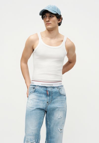 Νέος άνδρας που φοράει ένα μπλε φθαρμένο καπέλο, λευκό αμάνικο μπλουζάκι και ανοιχτό μπλε σκισμένα τζιν, στέκεται μπροστά σε ένα απλό φωτεινό φόντο.