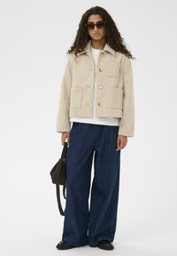 Veste cropped beige avec quatre poches avant et fermeture à boutons. Associée à un pantalon en denim foncé à jambes larges et un petit sac marron.