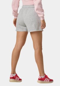 Person iført lysegrå shorts med elastik i taljen, lyserød sweatshirt og røde Adidas-sneakers, stående med ryggen til på hvid baggrund.