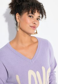 Maglione a V in lavanda con grafica "LOVE" beige, abbinato a grandi orecchini a cerchio dorati. Morbido tessuto a maglia, vestibilità casual.