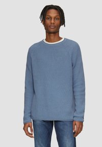 Pull en maille bleu avec un col rond et des poignets côtelés, associé à un jeans bleu sur un fond neutre.