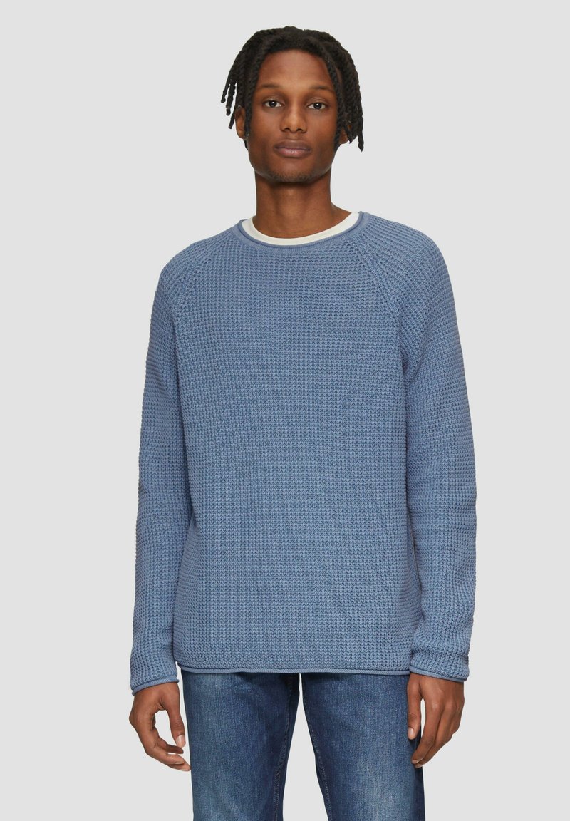 Pull en maille bleu avec un col rond et des poignets côtelés, associé à un jeans bleu sur un fond neutre.
