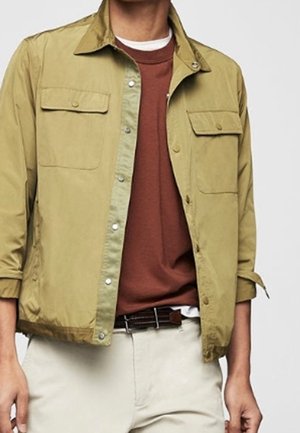 Torso maschile che indossa una giacca beige con bottoni sopra una maglietta color ruggine e pantaloni beige chiaro con una cintura marrone scuro.