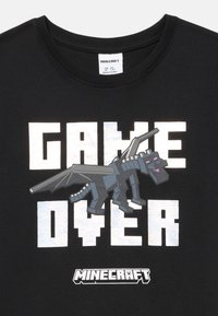 Czarna koszulka Minecraft z białym napisem "GAME OVER" oraz pixelizowanym czarnym graficznym smokiem umieszczonym centralnie nad logo Minecraft.