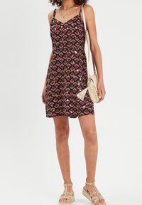 Femme portant une robe sans manches noire à motifs avec des bretelles fines, des sandales compensées beiges et un sac bandoulière tissé de couleur crème.