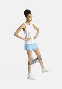 Top sin mangas ligero blanco y pantalones cortos azules. Banda de ejercicio de goma alrededor de los muslos. Zapatillas deportivas blancas. Posición de pie con las manos en las caderas.