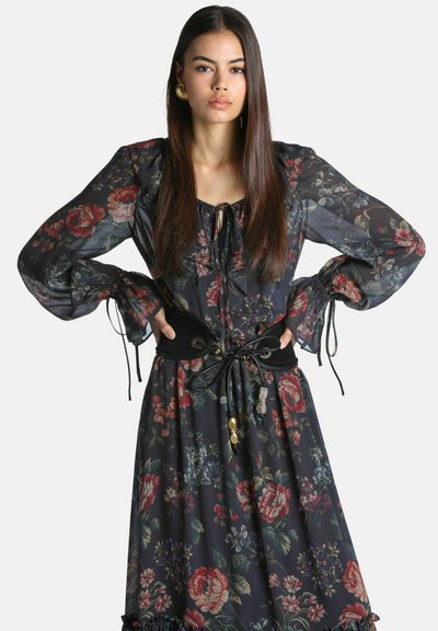 Femme aux longs cheveux foncés portant une robe noire à fleurs avec des manches bouffantes et une large ceinture noire nouée à la taille.