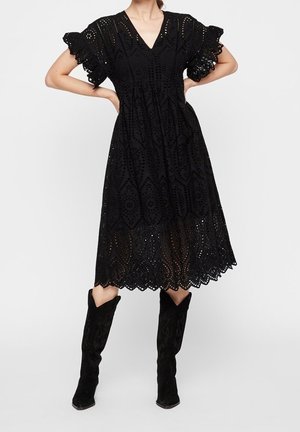 Femme debout, les mains sur les hanches, portant une robe midi noire en broderie anglaise avec des manches volantées et des bottes noires montantes jusqu'au genou.