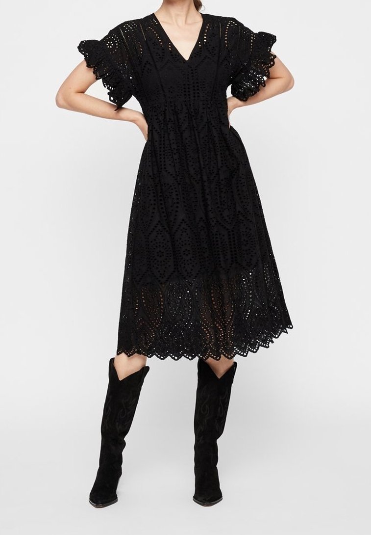 Femme debout, les mains sur les hanches, portant une robe midi noire en broderie anglaise avec des manches volantées et des bottes noires montantes jusqu'au genou.