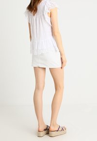 Blusa bianca strutturata con dettagli in pizzo, abbinata a una gonna in denim bianca aderente. La modella indossa sandali con zeppa beige con dettagli blu.
