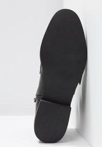 Semelle de botte noire avec talon texturé et avant lisse, taille 4 indiquée, positionnée verticalement contre un mur d'angle blanc.