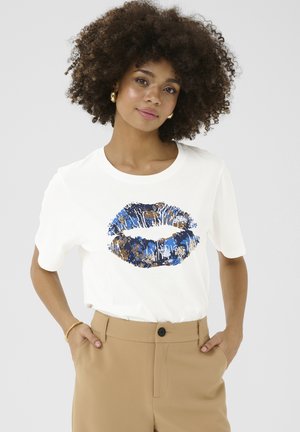 Camiseta de algodón blanca con un diseño de estampado de labios en azul y dorado. Manga corta, escote redondo y corte holgado, combinada con pantalones beige.