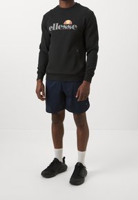Felpa nera con il logo "ellesse", tasca con zip e polsini a costine abbinata a pantaloni corti blu e scarpe da ginnastica nere. Calzini bianchi visibili.
