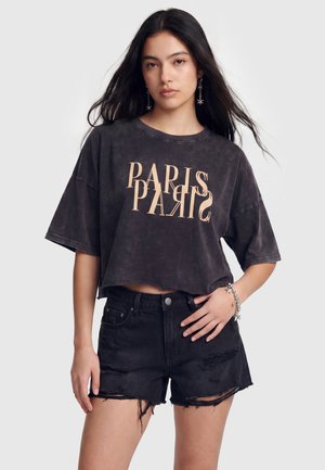 Młoda kobieta z długimi ciemnymi włosami, ubrana w czarny krótki T-shirt z napisem "PARIS" oraz czarne podniszczone jeansowe szorty, stojąca na jednolitym tle.