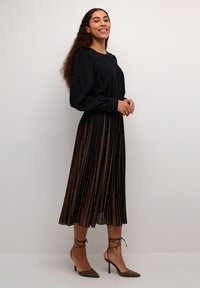 Schwarzes Langarmkleid mit einem gefalteten braunen Rock, Rundhalsausschnitt und geknöpften Ärmeln. Kombiniert mit braunen Absatzschuhen.
