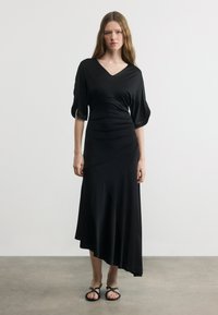 Γυναίκα που φοράει μαύρο φόρεμα με V-λαιμόκοψη, μανίκια μέχρι τον αγκώνα και ασύμμετρο τελείωμα, και μαύρα σανδάλια, σε απλό φόντο.