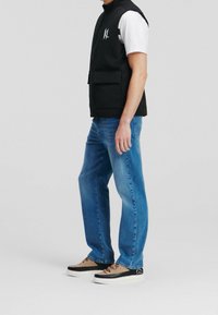 Svart ärmlös väst med framfickor över en vit t-shirt, i kombination med ljusblå jeans och flerfärgade sneakers. Neutral bakgrund.