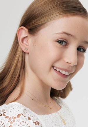 Złote kolczyki w kształcie kwiatu noszone przez dziewczynę z długimi włosami, w połączeniu z warstwowymi, delikatnymi złotymi naszyjnikami i białą koronkową bluzką.