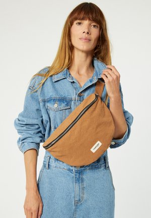 Braune Stoff-Schultertasche mit geschwungener Form und schwarzem Reißverschluss, ausgestattet mit einem Logo-Anhänger und quer über der Brust getragen, kombiniert mit einer Jeansjacke.