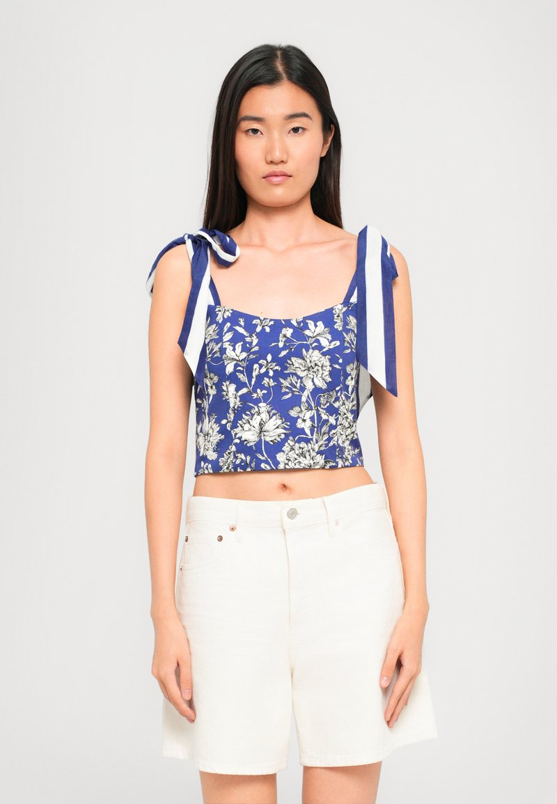 Woman wearing blue floral crop top with tied shoulder straps and high-waist white shorts, standing against plain light background.