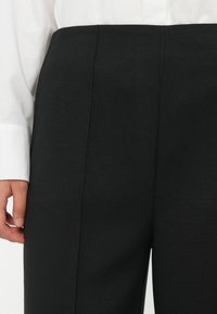 Close-up of a person wearing high-waisted black trousers and a white long-sleeve shirt, with their left hand visible by their side.