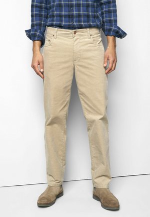 Mann trägt beige Cordhose, braune Wildlederschuhe und ein blau kariertes Langarmhemd mit hochgekrempelten Ärmeln, steht vor weißem Hintergrund.