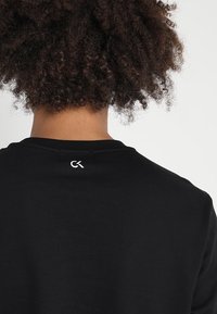 Sweatshirt en coton noir avec un col rond simple et un logo CK blanc au-dessus de la zone du cou, présentant une texture lisse.