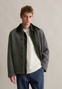 Geruite flanellen jas in zwart en grijs, met een corduroy kraag, knoopsluiting en twee voorzakken. Draagt over een crèmekleurig T-shirt.