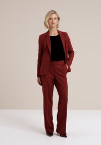 Žena stojí s dôverou v priliehavom burgundskom obleku s čiernym topom, predvádzajúc modernú obchodnú módu na neutrálnom pozadí.