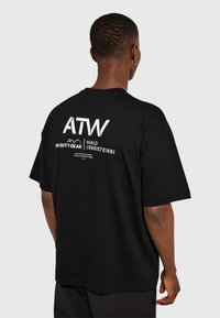 Mężczyzna o krótkich włosach i kolczyku, ubrany w czarną, oversize'ową koszulkę z białym napisem i logo na plecach, na jednolitym tle.