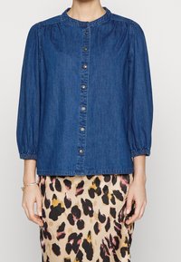 Donna che indossa una camicia di jeans blu con bottoni e maniche a 3/4 arricciate, insieme a una gonna beige con stampa leopardata.