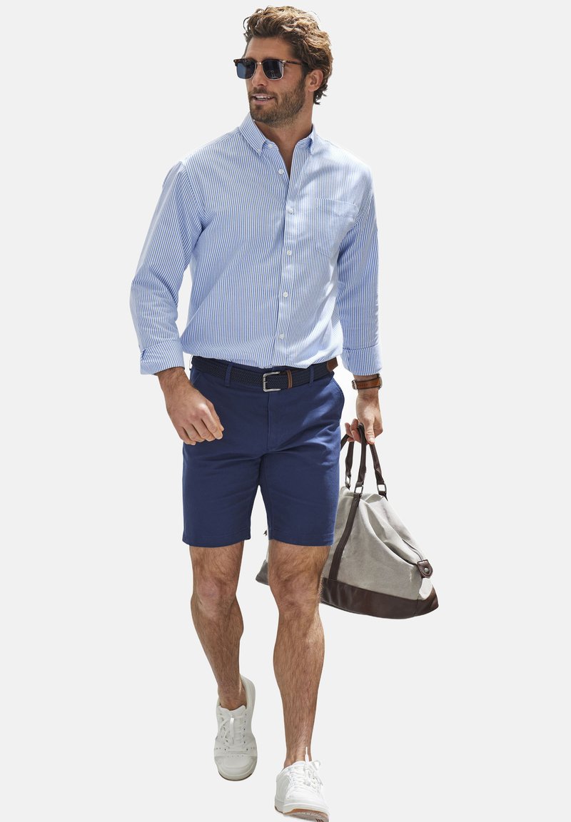 Hellblau und weiß gestreiftes Hemd mit Knöpfen, marineblaue Shorts, weiße Turnschuhe und eine grau-braune Sporttasche tragend.