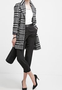 Černobílý dlouhý kabát s vzorem pepito přes bílý top, doplněný černými zkrácenými kalhotami a černými lodičkami na podpatku. Součástí je černá kabelka.