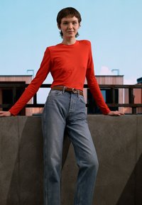 Camisa vermelha de mangas compridas combinada com jeans de brim azul claro de cintura alta, com um cinto castanho e um fundo de betão liso.