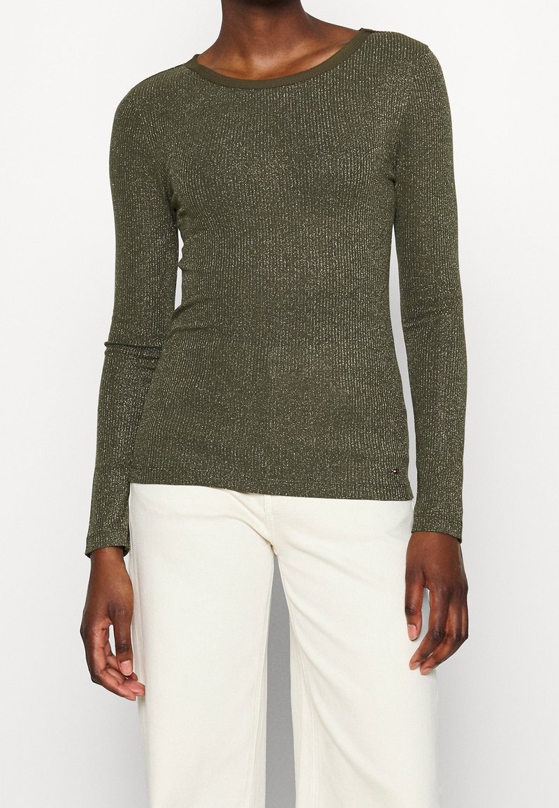 Olijfgroene top met lange mouwen, ribbeltextuur, ronde halslijn en subtiele metalen accenten. Slanke pasvorm, gedragen met lichtgekleurde broek.