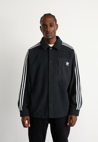Černá fleecová košile s výstřihy na knoflíky, bílými pruhovanými rukávy a kapsou na levé straně hrudi, která nese logo Adidas.