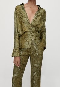 Blouse en satin vert olive avec imprimé serpent, présentant un décolleté en V plongeant, une taille ceinturée et une coupe décontractée. Manches longues et poignets repliés.